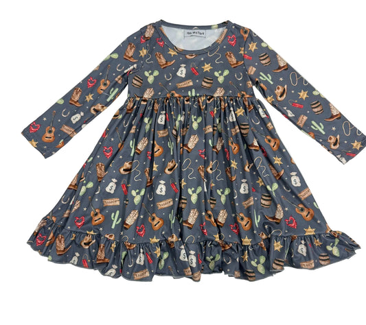 A long-sleeve children's dress with a gathered waist and ruffled hem, featuring a western-themed print of cacti, cowboy boots, lasso ropes, stars, and hats on a slate blue-gray background.