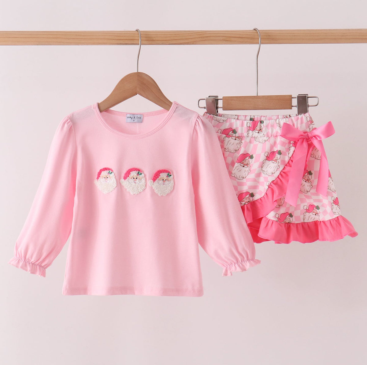 A children's two-piece outfit hanging on wooden hangers against a light pink wall. The set includes a long-sleeve pink top with three small Santa patches on the chest and matching ruffled shorts with a floral print, pink bow, and pink ruffle trim.